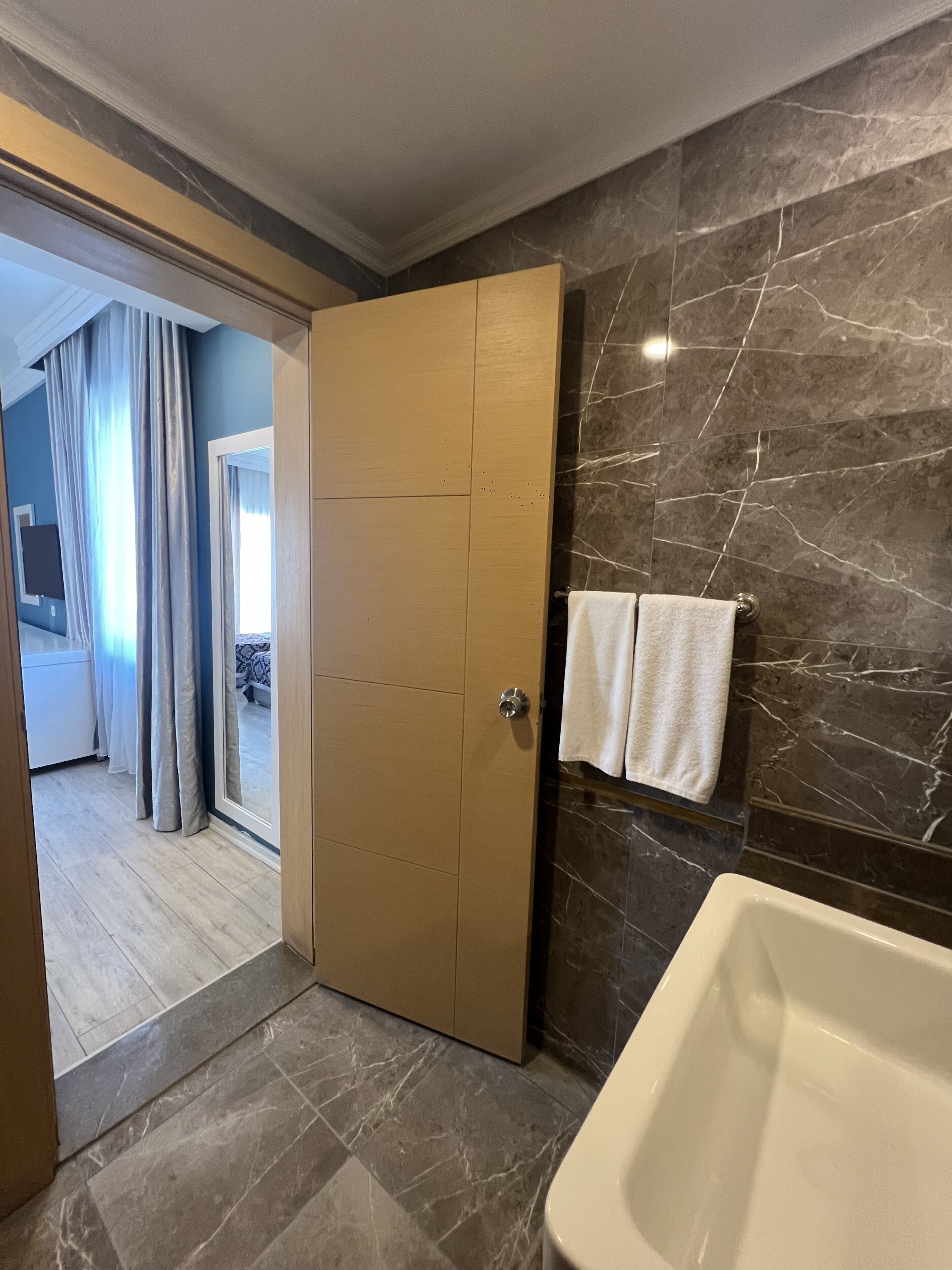Un baño moderno con pared de mármol, bañera y puerta de madera que da a un dormitorio.