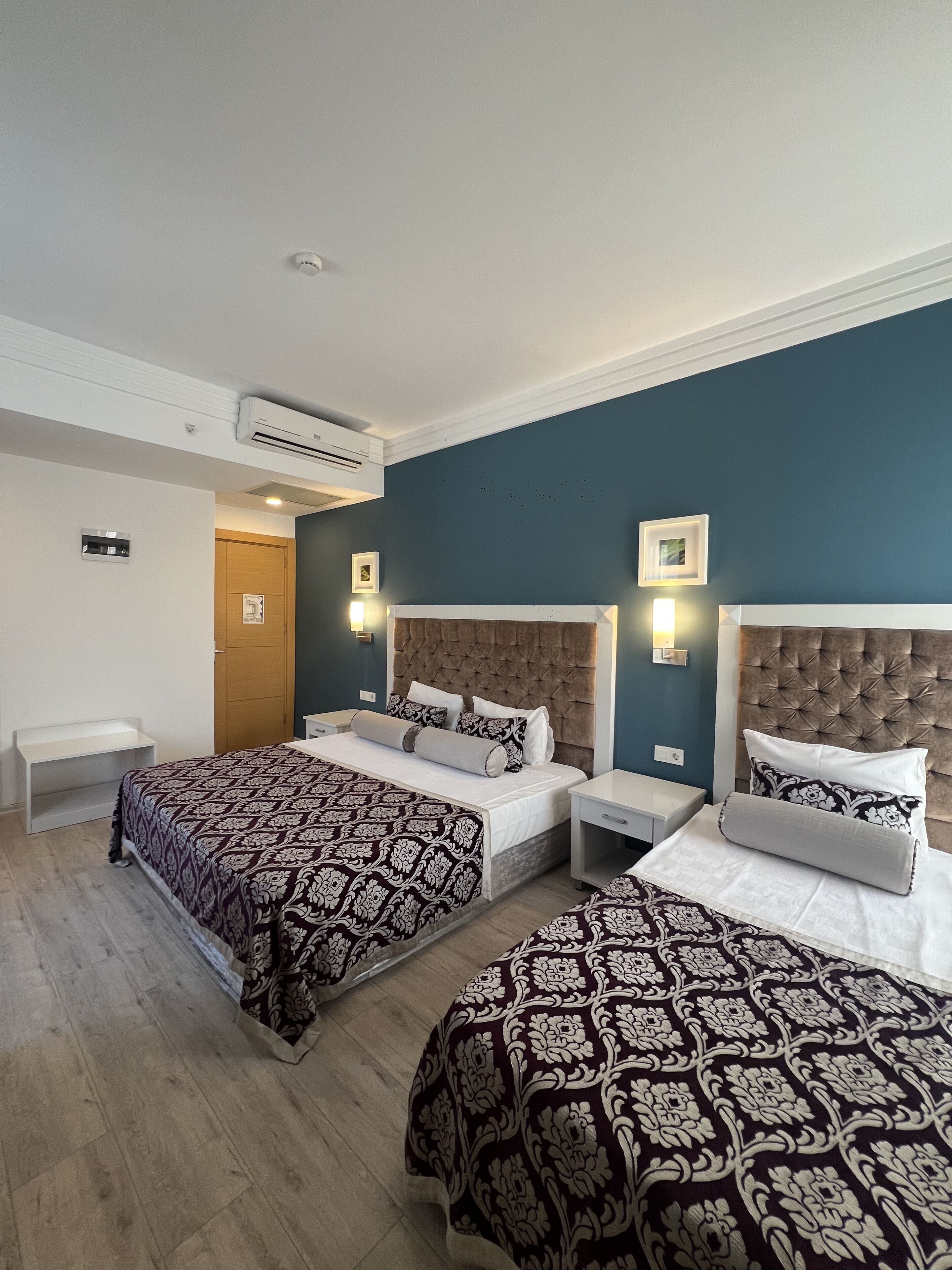 A hotel room with two beds, a wooden door, a white shelf, and a wall-mounted air conditioner.