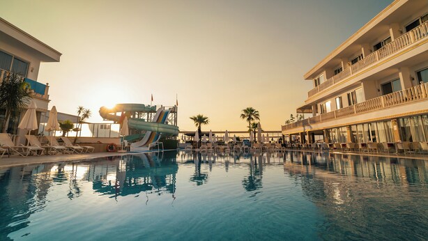 Una piscina con sillones de descanso y sombrillas, un tobogán acuático y un edificio con balcones.
