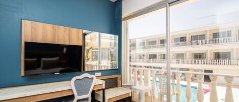 A room with a TV mounted on a wooden panel, a desk with a chair, and a large window overlooking a pool and a multi-story building.