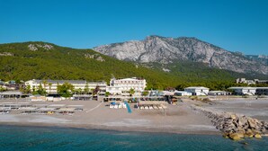 Un hotel costero con varias construcciones, una playa con tumbonas y una montaña al fondo.