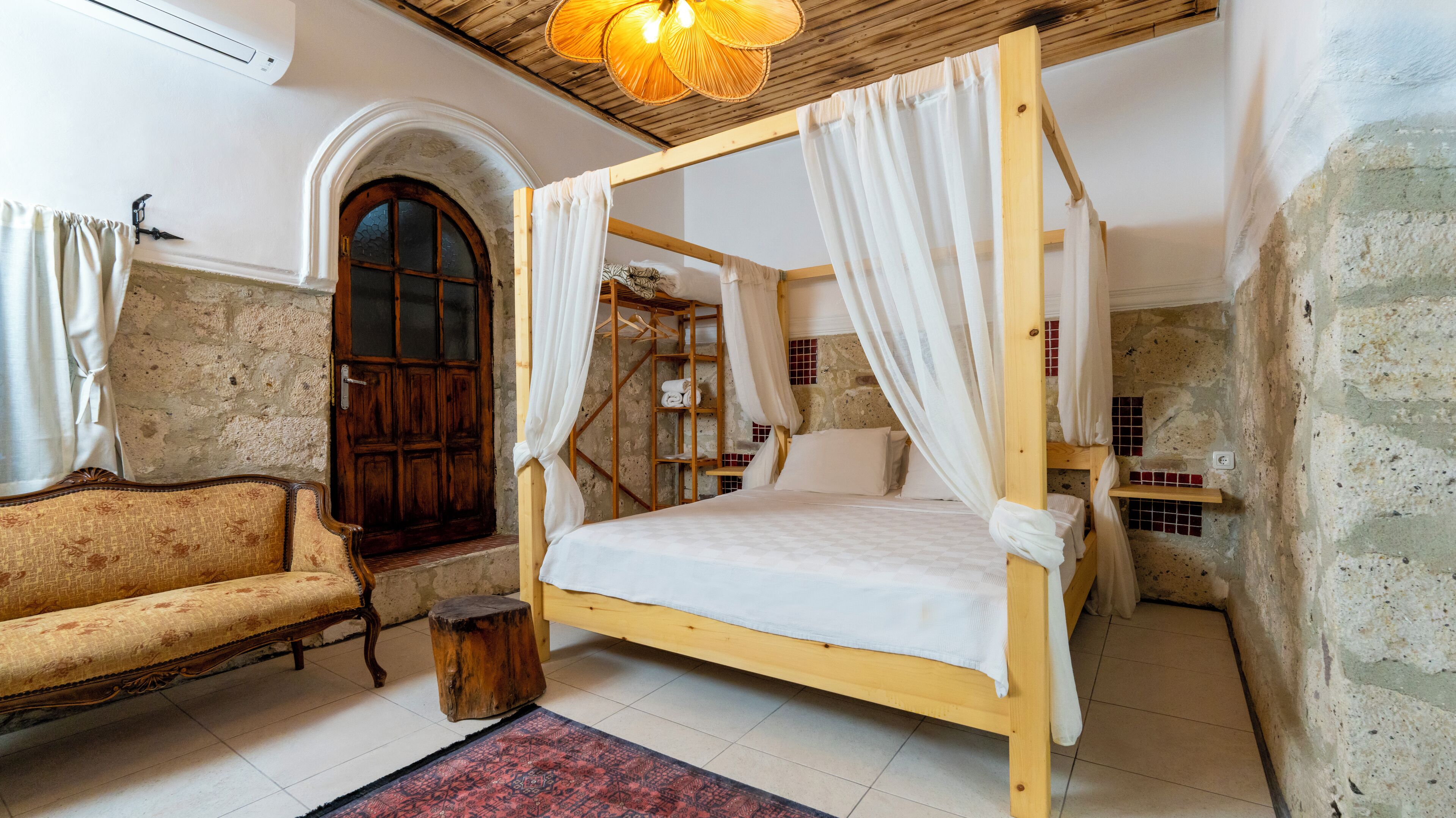 Una habitación rústica con cama con dosel, un banco de madera, una pared de piedra y una puerta de madera.