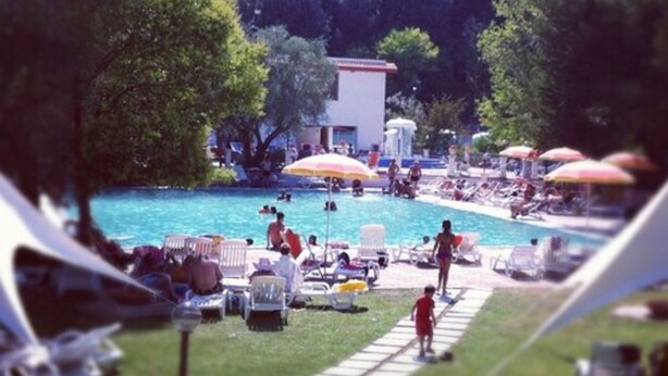 Area piscina con lettini e ombrelloni, circondata da alberi e un edificio sullo sfondo.
