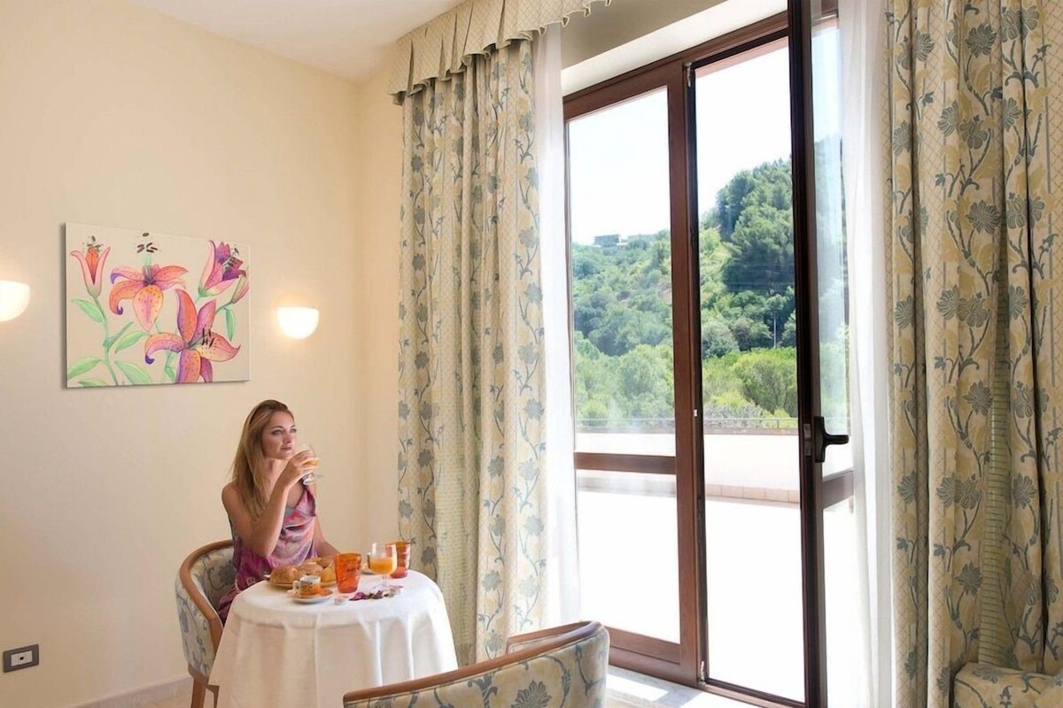 A dining area with a table set for two, a chair, a painting on the wall, and a view through a large sliding glass door.