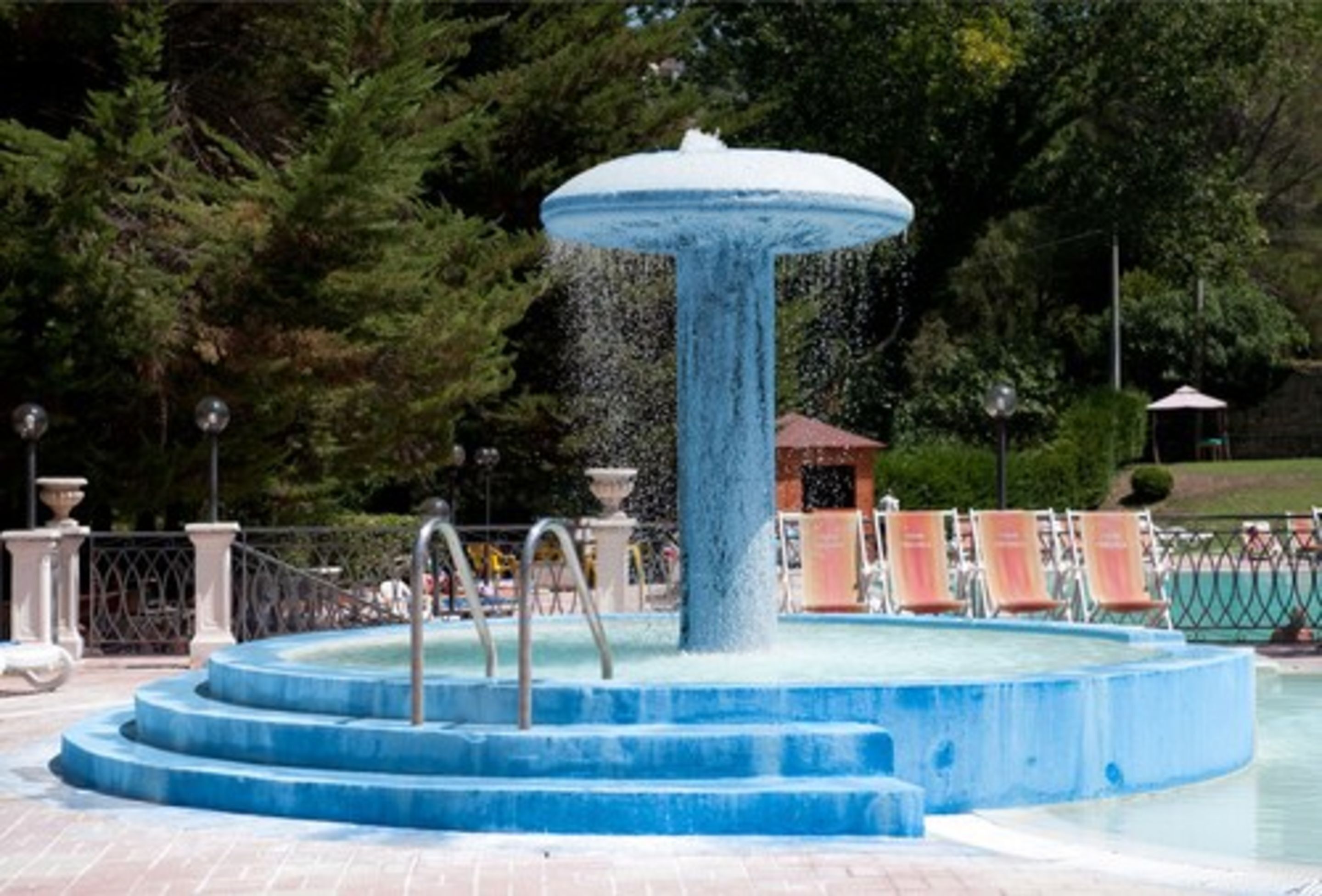 Ein Springbrunnen mit kreisförmigem Becken und Stufen, umgeben von einem Zaun und Bäumen.