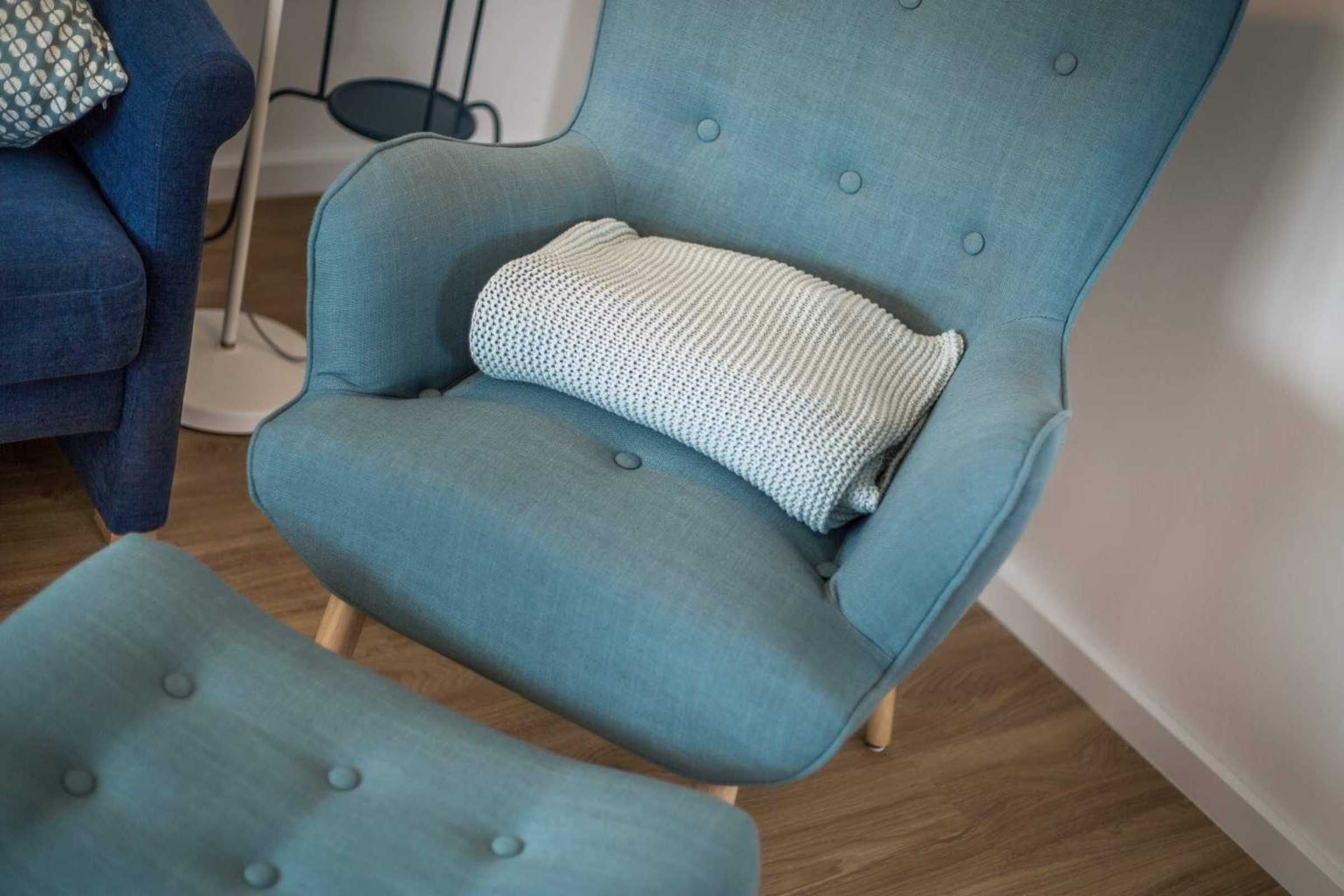 Un fauteuil bleu clair recouvert d’un tissu d’ameublement, accompagné d’un pouf assorti, d’un coussin blanc tricoté et d’un lampadaire en arrière-plan.