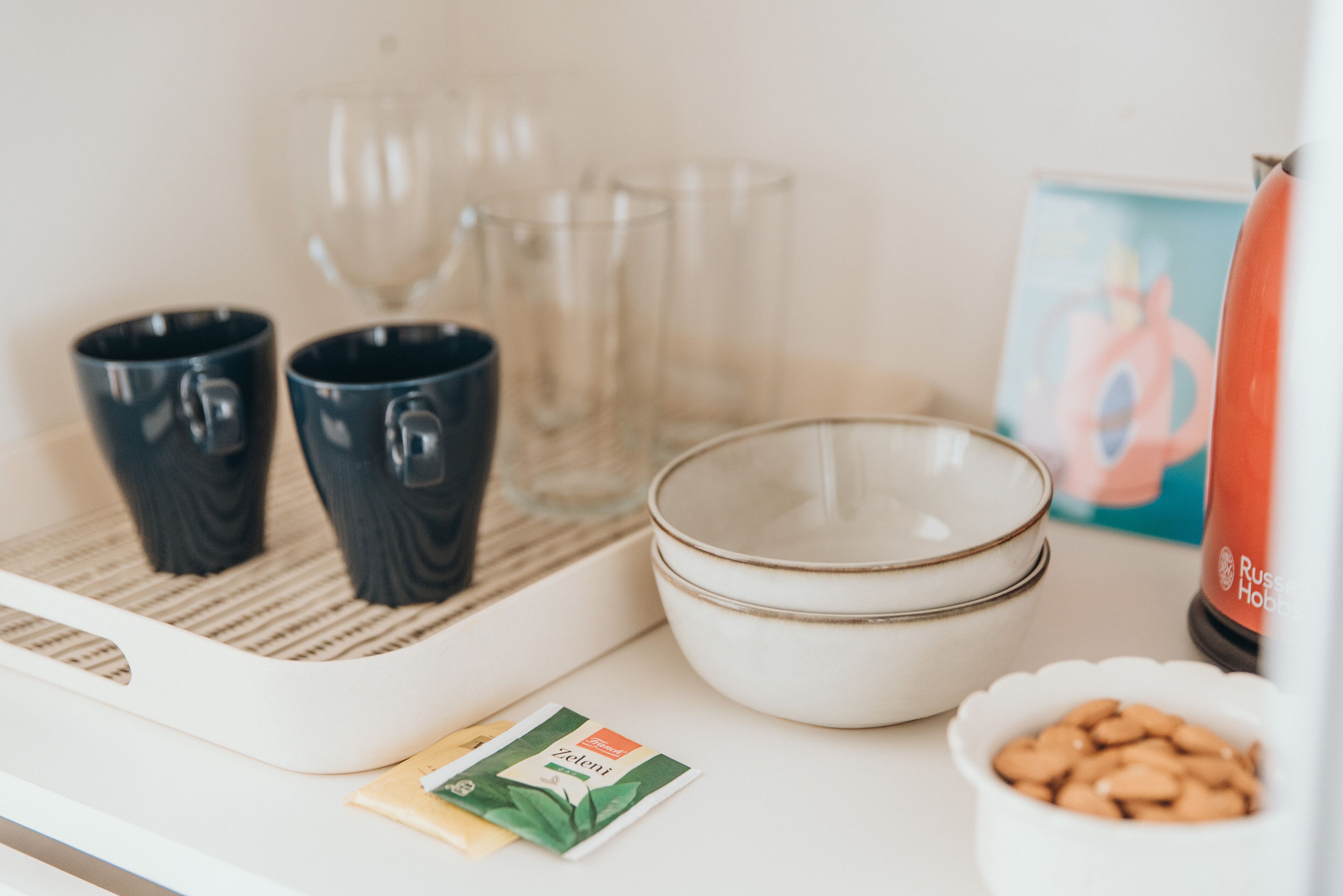 A tray with two black mugs, a white bowl, a small bowl of almonds, a packet of tea, and a bottle of hot water.