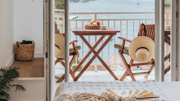 Un dormitorio con cama, suelo de madera, planta en maceta, cesta de mimbre y vista al mar por una puerta abierta.