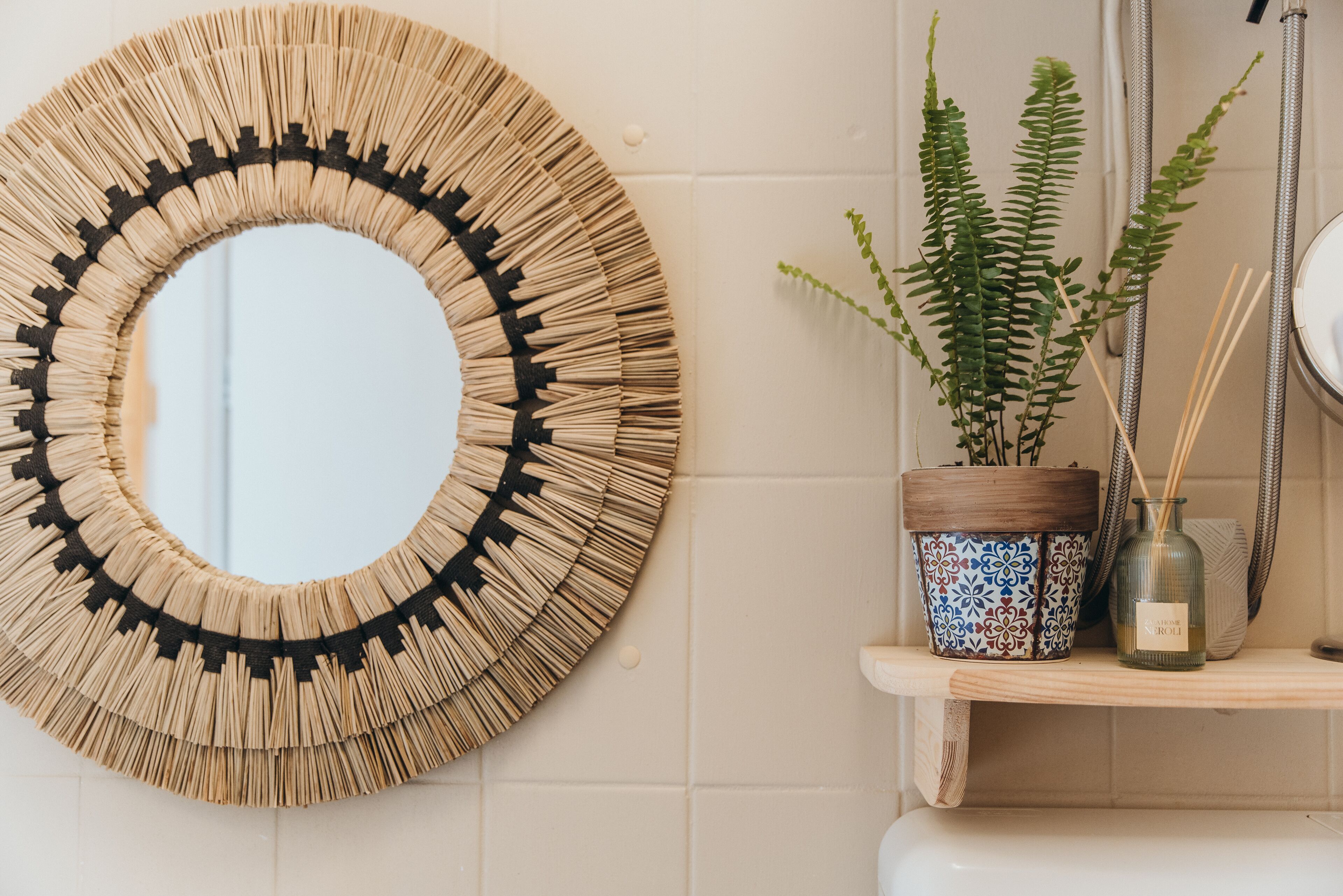A round mirror with a woven frame, a potted fern, and a reed diffuser on a shelf.