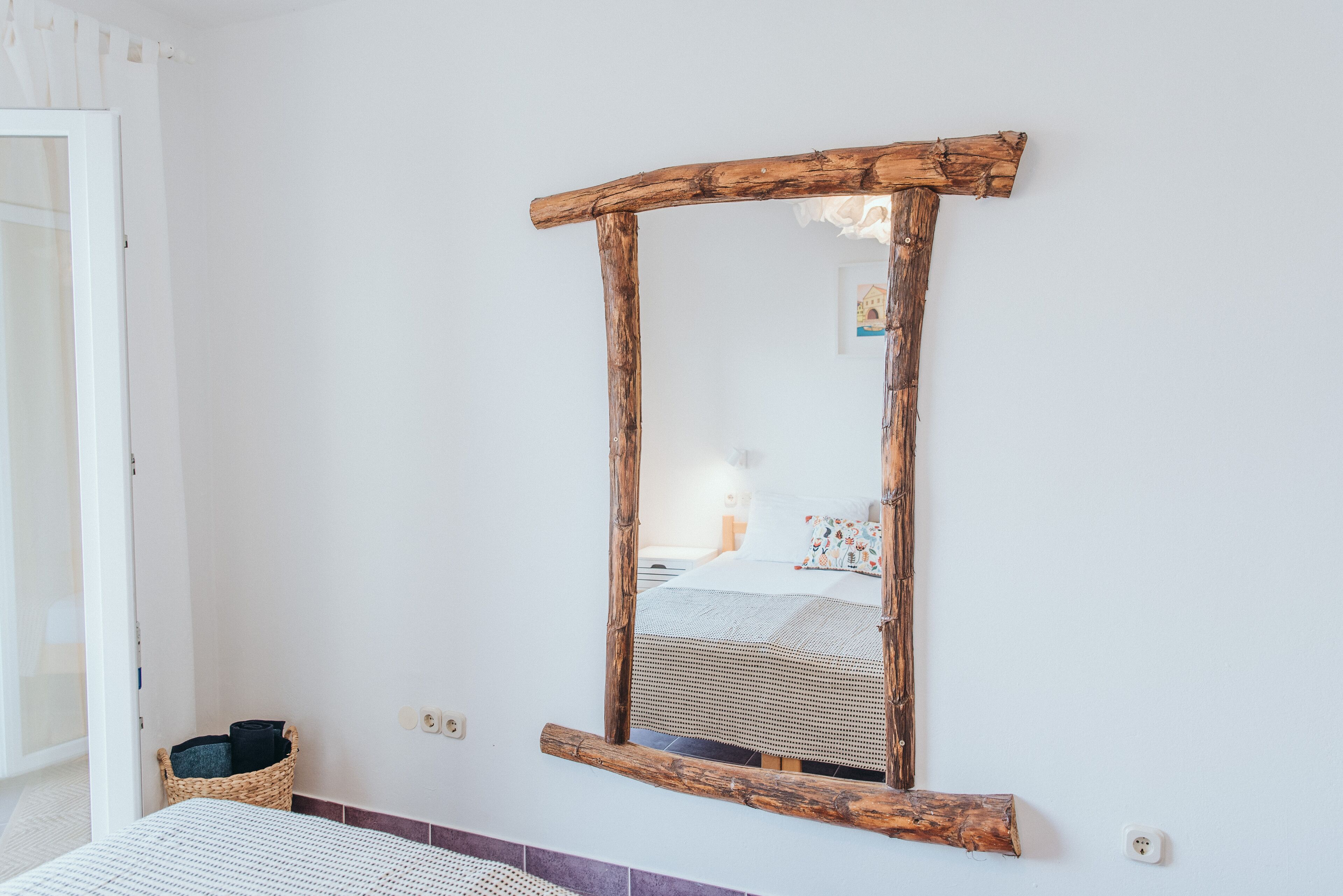 A bedroom with a wooden-framed mirror, a bed with a white and patterned bedspread, and a wicker basket on the floor.