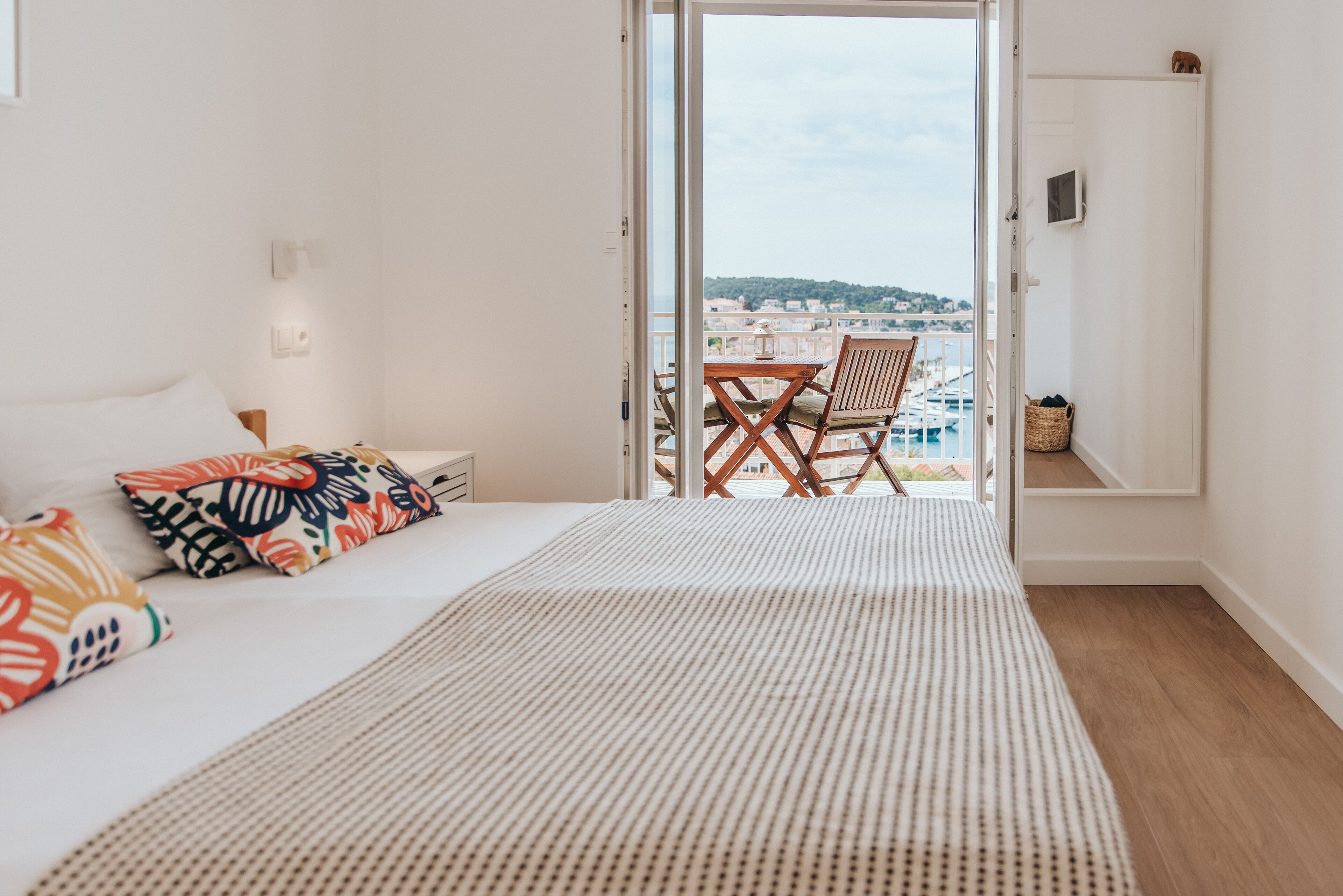 Een slaapkamer met een bed, kussens, een nachtkastje, een raam en een balkon met uitzicht op zee en gebouwen.