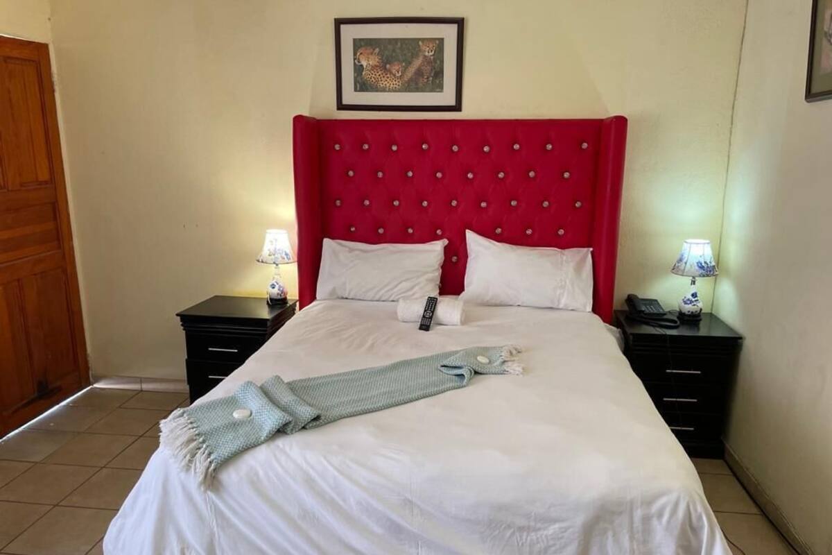 Un lit bien fait, avec une tête de lit rouge, une literie blanche et un plaid bleu clair. Il y a deux tables de chevet avec des lampes et un tableau encadré accroché au mur au-dessus du lit.