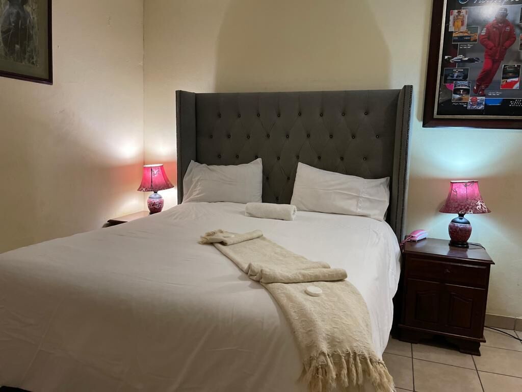 Een net opgemaakt bed met een grijs hoofdbord, witte kussens en een beige plaid. Er staan twee nachtkastjes met lampen en een klein decoratief object op één.