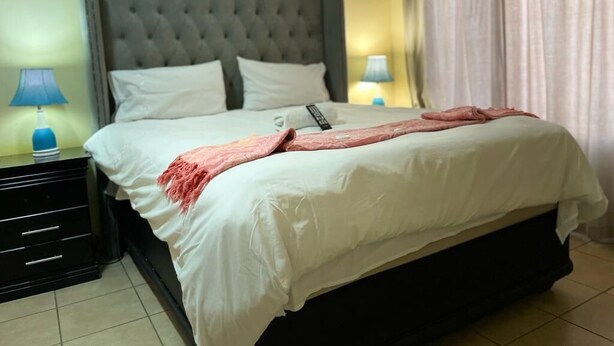A neatly made bed with white linens and a pink throw blanket, a grey tufted headboard, and a nightstand with a lamp.