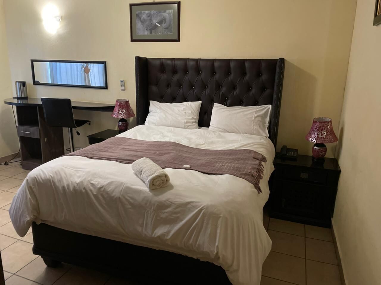 A neatly made bed with a rolled towel, a television mounted on the wall, a desk with a chair, and a nightstand with a lamp.