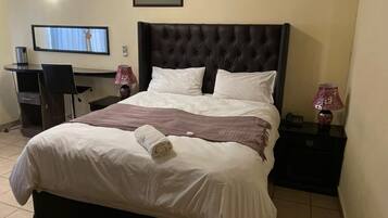 A neatly made bed with a rolled towel, a television mounted on the wall, a desk with a chair, and a nightstand with a lamp.
