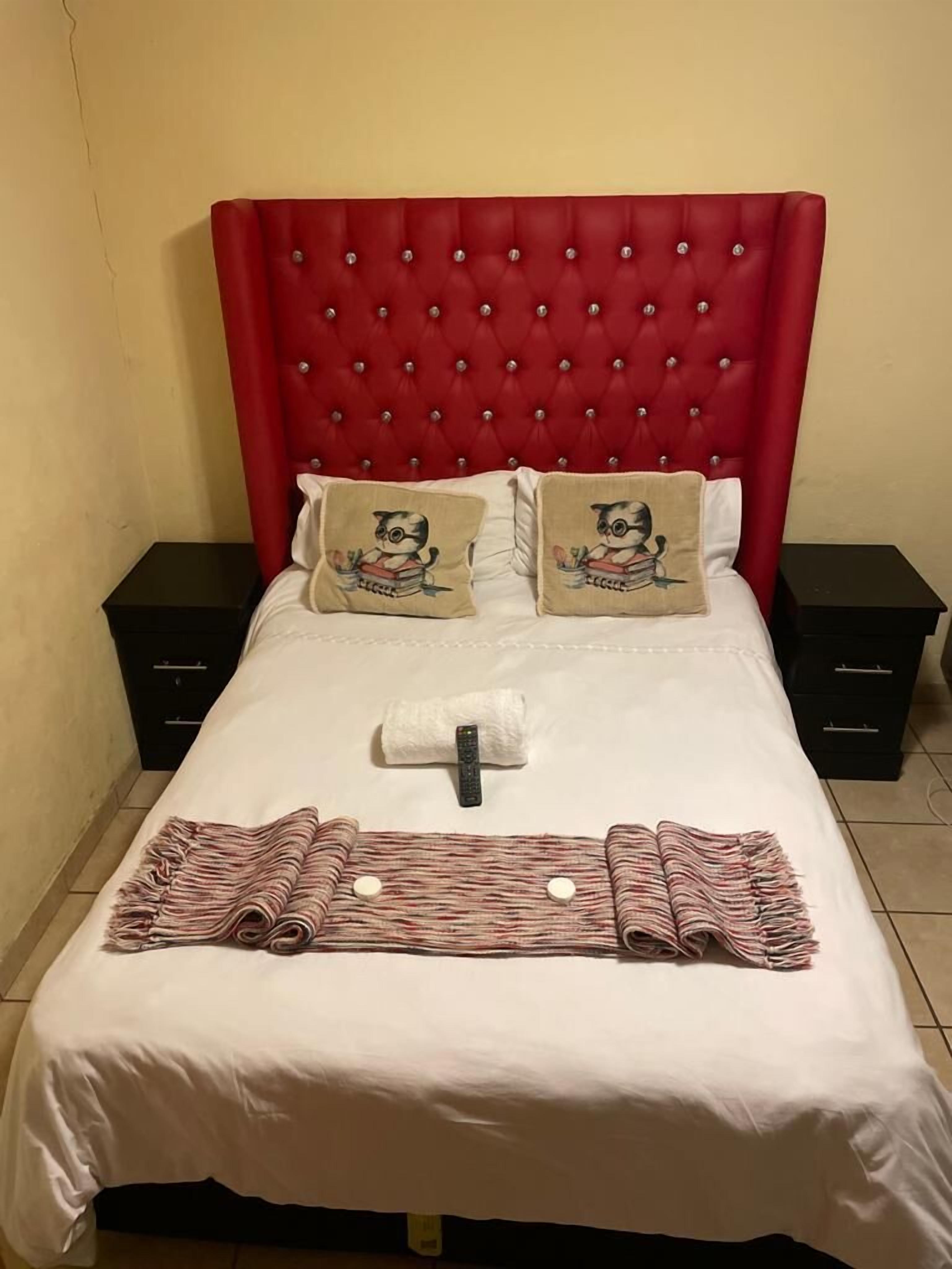 A neatly made bed with a red tufted headboard, two pillows with a cat design, and a folded towel on the bed.
