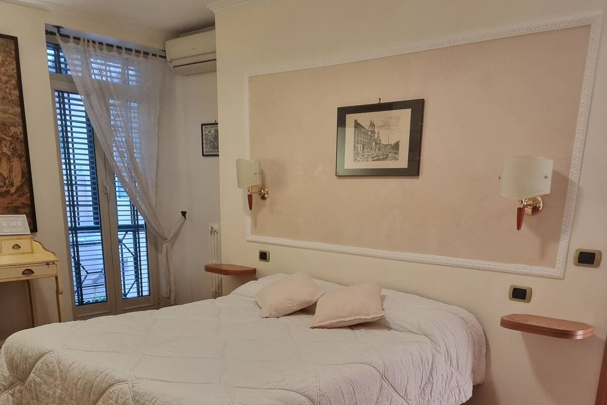 A bedroom with a large bed, wooden flooring, a nightstand, a bedside table, and a window with curtains.