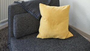 A modern living room with a grey sofa, a yellow checkered pillow, a green curtain, and a wall-mounted shelf with books. - Apartments 4 YOU - Lange Strasse (Fuerth)