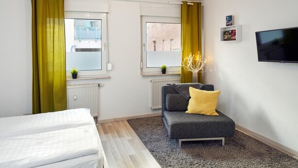 A hotel room with two beds, a grey sofa with a yellow pillow, a TV on the wall, and windows with curtains. - Apartments 4 YOU - Lange Strasse (Fuerth)