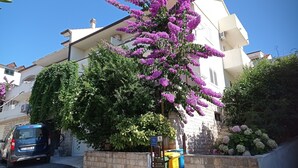 A building with a stone wall, a parked car, and a tree with purple flowers. - Apartments and Rooms Zvjezdana (Hvar)