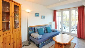 A cozy living room with a wooden cabinet, a blue sofa with cushions, a wooden table, and a large window with red curtains. - Apartment 8 Strandstar (ID 118) (Sankt Peter-Ording)