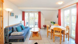 A cozy living room with a sofa, a dining table, and chairs. There is a TV on the wall and a window with curtains. - Apartment 8 Strandstar (ID 118) (Sankt Peter-Ording)