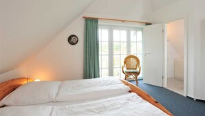 A bedroom with a sloped ceiling, a bed with white linens, a wicker chair, and a window with green curtains. - Apartment 8 Strandstar (ID 118) (Sankt Peter-Ording)