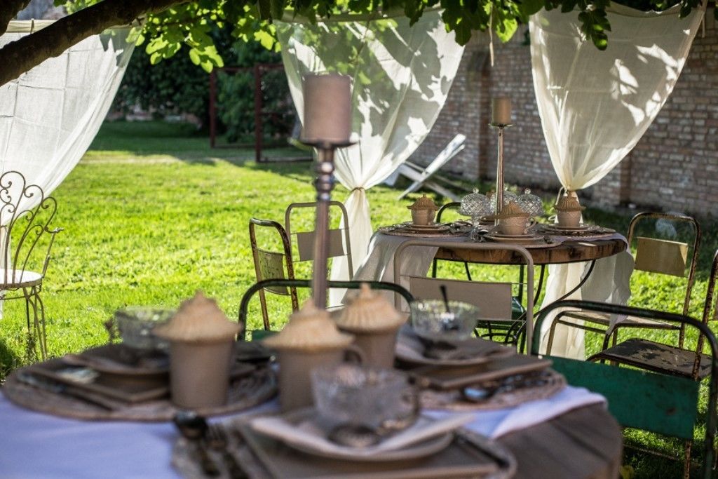 An outdoor dining area with tables set under a tree, surrounded by chairs and covered with white cloths.