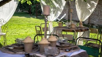 An outdoor dining area with tables set under a tree, surrounded by chairs and covered with white cloths.