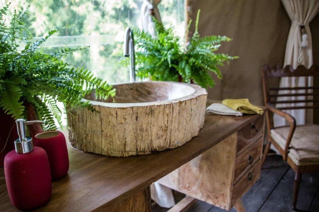 Una mesa de madera con un lavabo circular de madera, dispensadores de jabón rojo y un helecho en maceta.