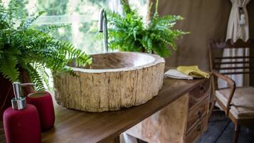 Una mesa de madera con un lavabo circular de madera, dispensadores de jabón rojo y un helecho en maceta.