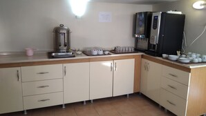 Cozinha compacta com máquina de café, chaleira e micro-ondas. Há armários com gavetas e uma bancada com diversos objetos.