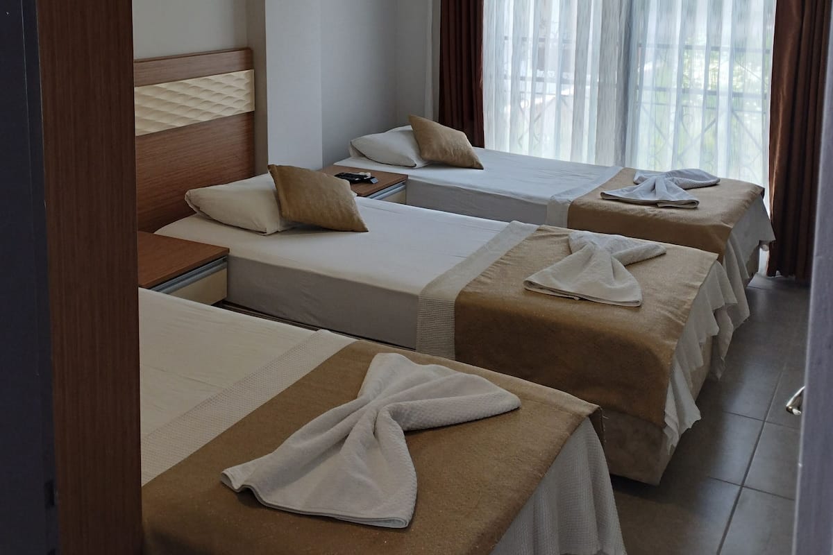 A hotel room with four single beds, each with a towel and a pillow. There is a wooden headboard on the left side, and a window with sheer curtains.
