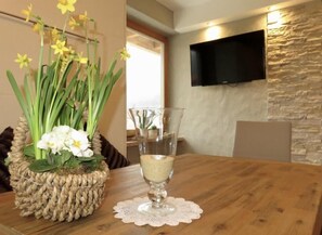 A dining table with a woven basket of flowers, a clear glass with a white substance, and a television mounted on a stone-textured wall.