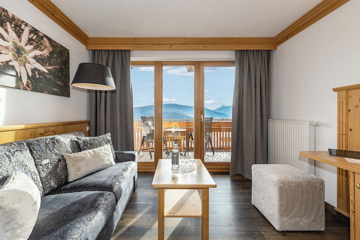 A cozy room with a sofa, a small table, a bench, and a view of mountains through large glass doors.