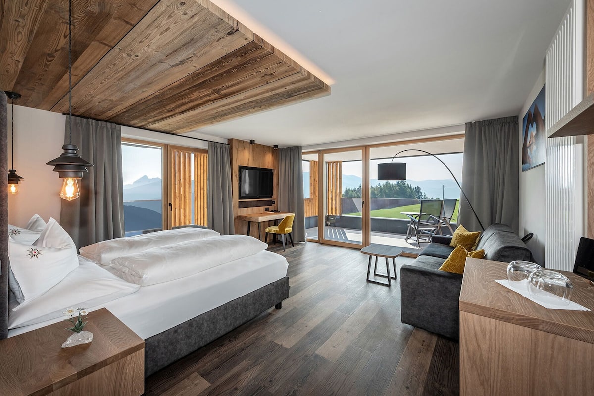 A modern hotel room with a large bed, a desk, and a balcony with a view of the mountains.