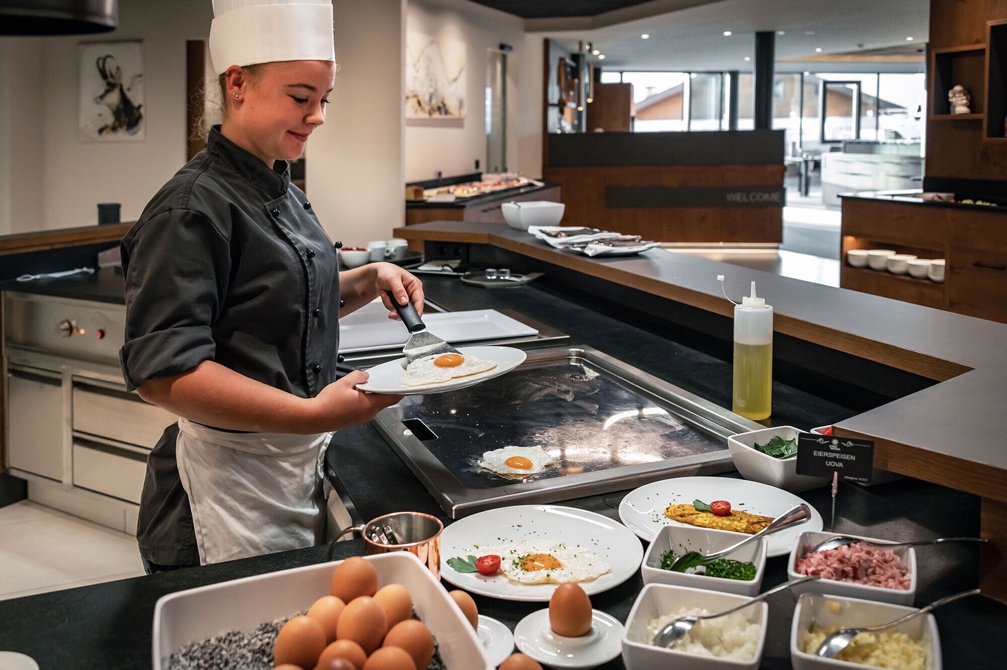 A chef preparing an egg on a hot griddle in a modern kitchen with various food items and condiments on the counter.