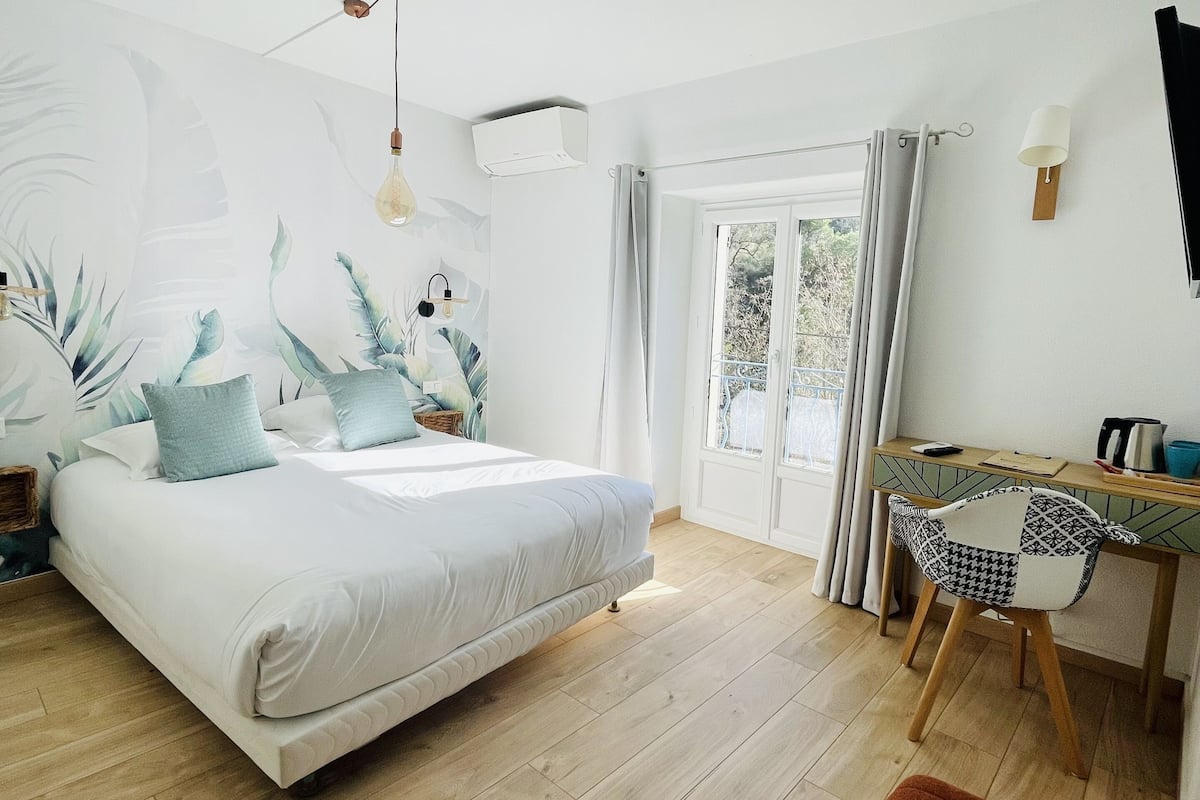 Een slaapkamer met een bed, een bureau en een stoel. Er is een raam met gordijnen, een wandairconditioner en een lamp.