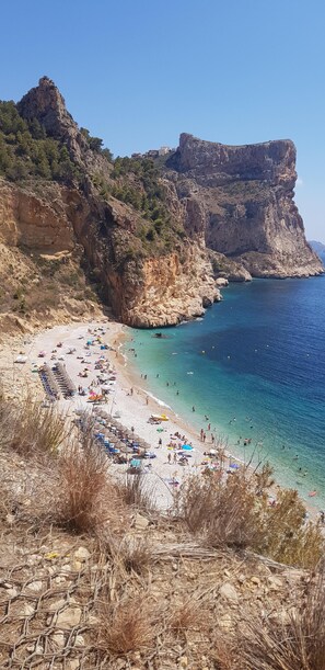 Una playa costera con aguas azul turquesa, acantilados rocosos y una orilla arenosa con sombrillas y gente.