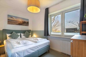 A neatly made bed with pillows, a bedside lamp, a window with a view of trees, and a painting on the wall.