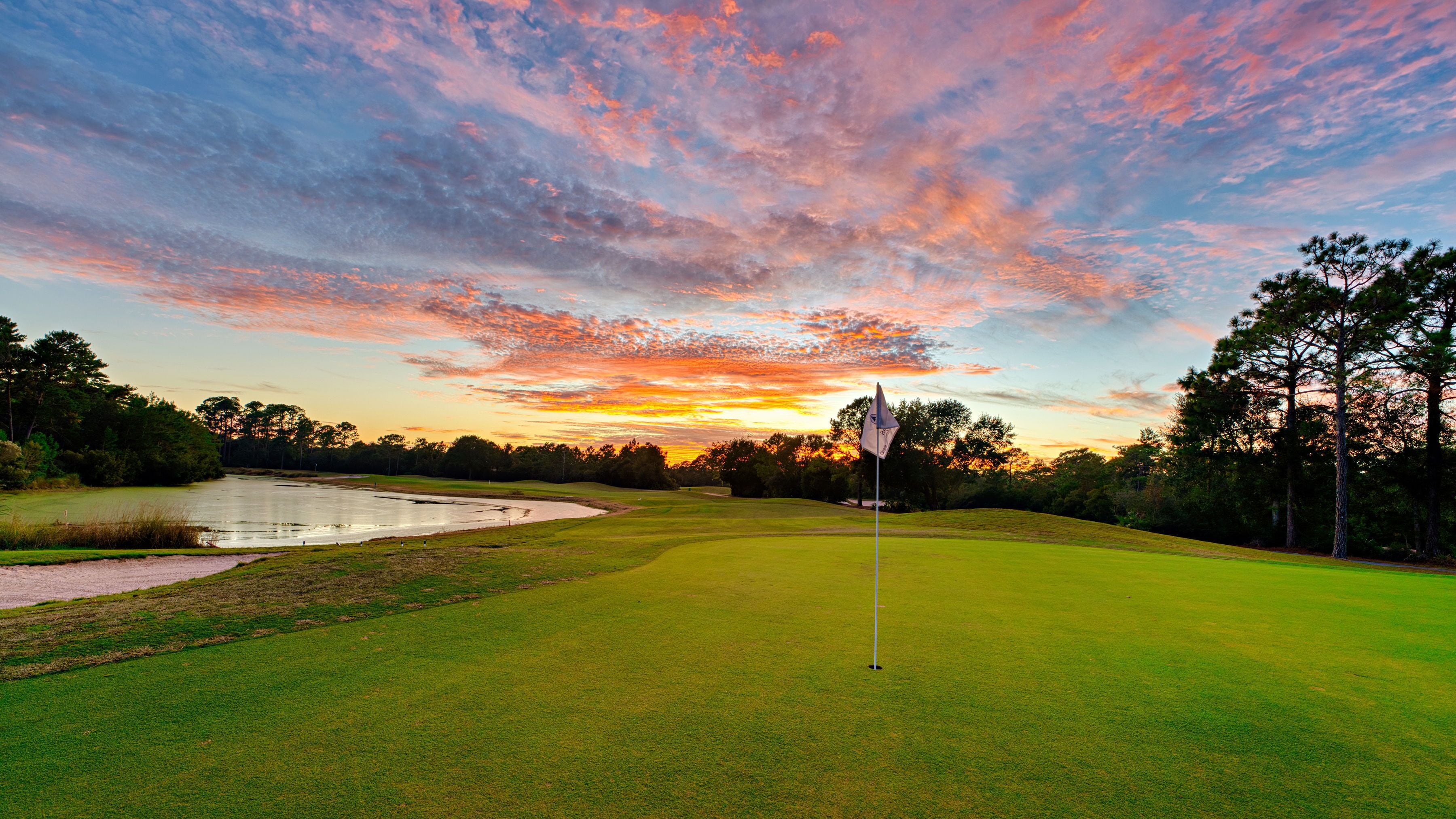 A golf course at sunset with a flagstick in the foreground, surrounded by trees and a body of water.