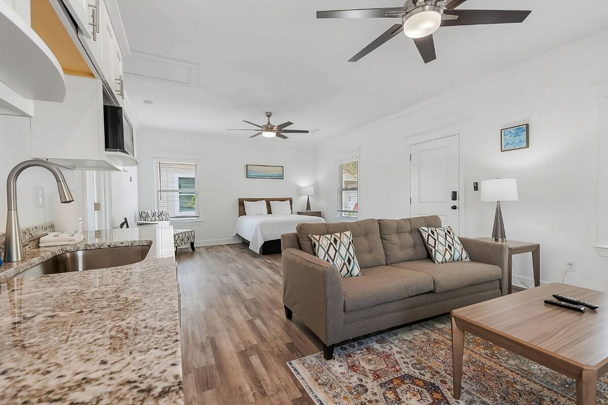 A modern living room with a sofa, a coffee table, a TV, a ceiling fan, and a kitchen area with a sink and countertop.