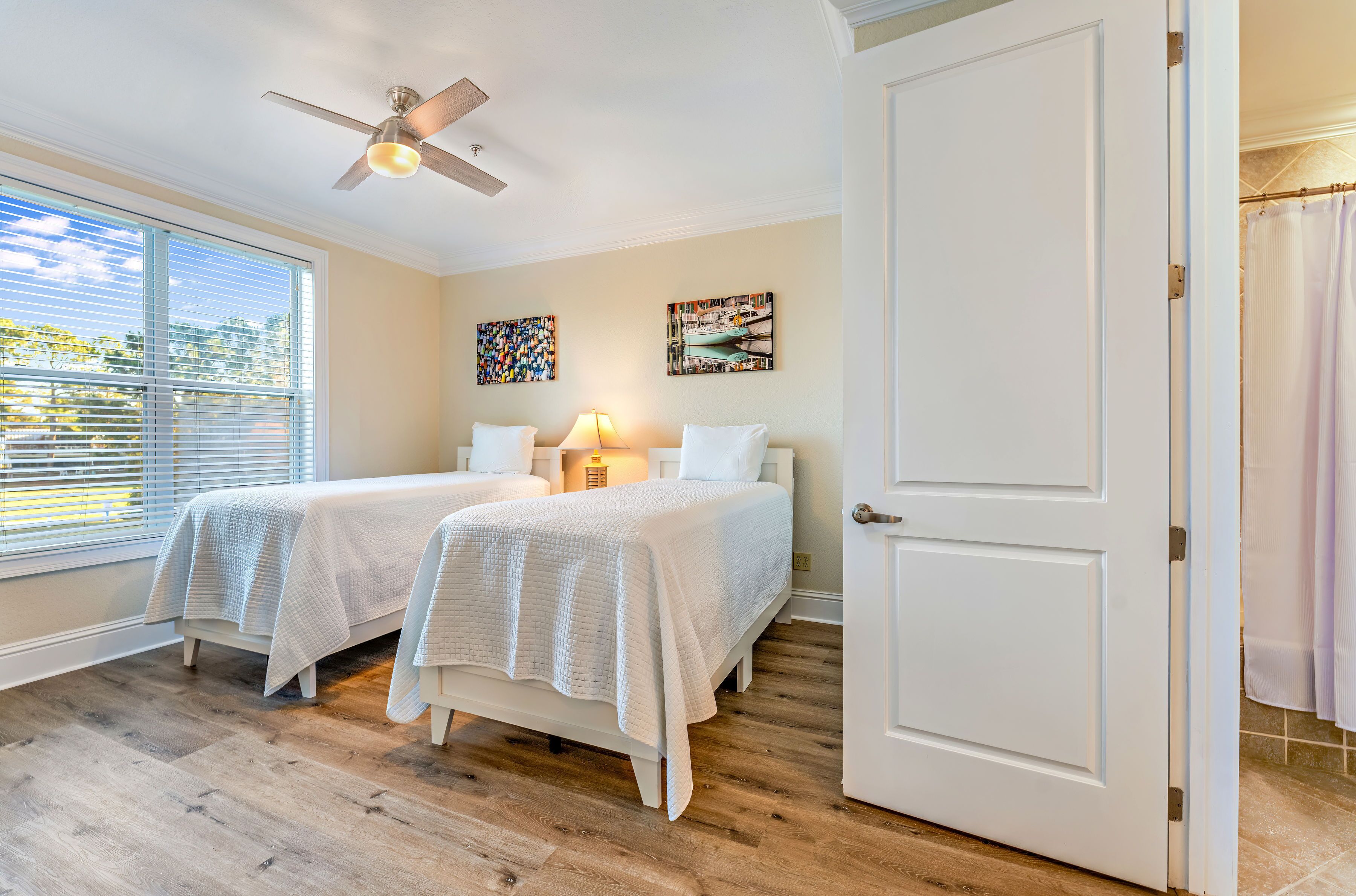 A bedroom with two beds, a ceiling fan, a window with blinds, and artwork on the wall.