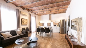 Una sala de estar moderna con piso de madera tipo espiga, un sofá marrón con cojines, una mesa de centro y un comedor con mesa y sillas de madera.