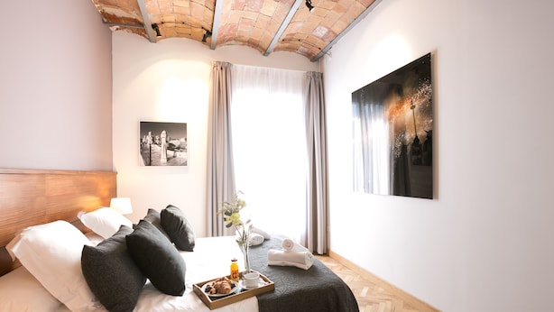 Habitación de hotel con cama, una bandeja con croissants y una bebida, un jarrón con flores y obras de arte en las paredes.