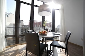 A dining area with a round table, black chairs, and a bowl of fruit. Large windows offer a view of buildings outside. - Serennia Apartments Ramblas-Pl.Catalunya (Barcelona)