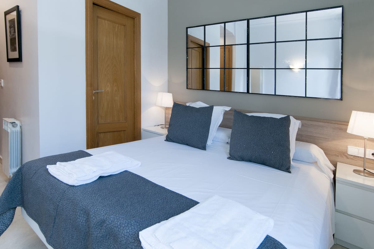A neatly made bed with a grey blanket, two pillows, and a white towel. There is a wooden door, a nightstand with a lamp, and a large mirror on the wall.