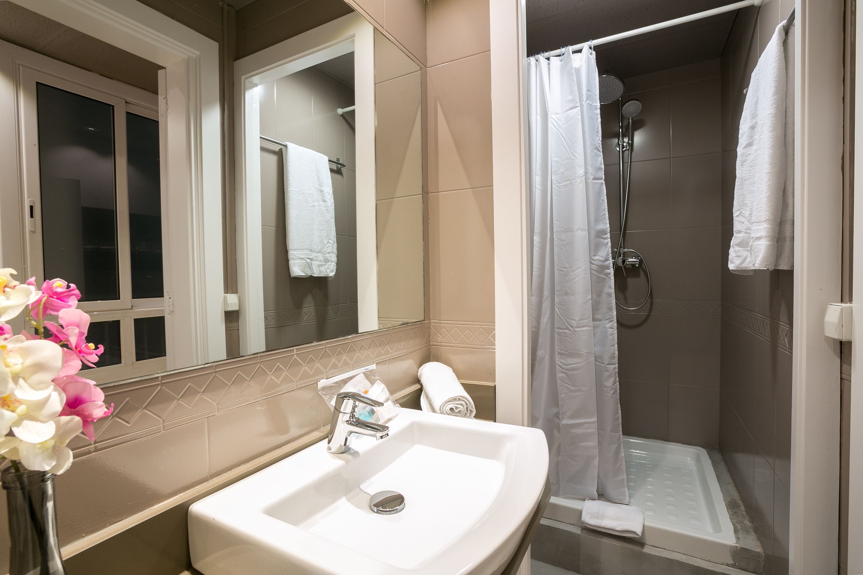 A bathroom with a sink, mirror, shower, and a window with a towel hanging.