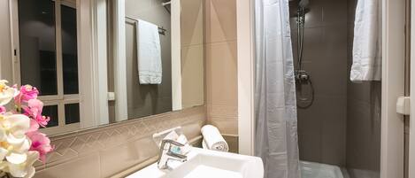 A bathroom with a sink, mirror, shower, and a window with a towel hanging.